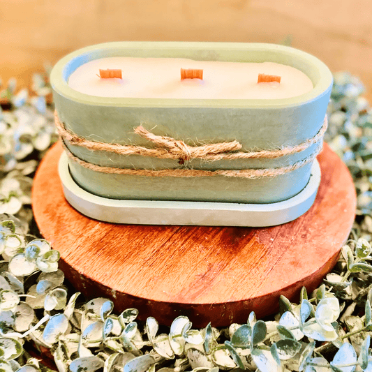 3 wooden wicks in soy wax inside a soft green cement vessel with a matching green tray. Sitting on a wooden circle on a bed of leaves. 
