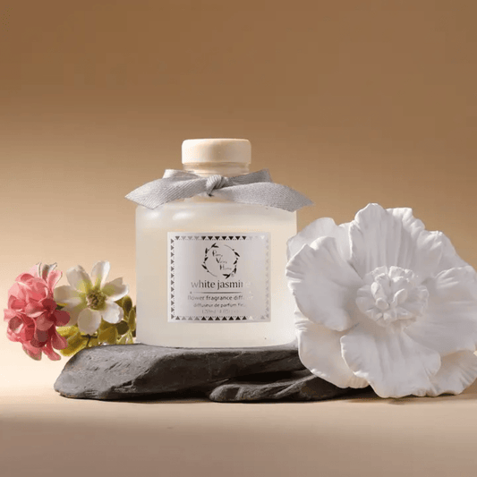 Frosted glass diffuser bottle shown placed on a flat rock. white ceramic open blossom flower laying next to the bottle, against a golden background. 